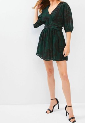Femme portant une robe courte verte et noire à motifs, taille froncée et manches bouffantes, associée à des sandales noires à talons hauts.