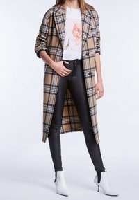 Manteau long beige et noir à carreaux, t-shirt blanc à motif floral, pantalon en cuir noir, et bottines blanches à bout pointu et petit talon.