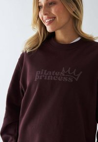 Bordowy sweter z logo "królewna pilates" w stylizowanej grafice z koroną. Wykonany z miękkiego materiału, ma luźny krój i okrągły dekolt.