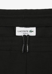 Pantalons noirs slim avec une taille à cordon. Présente une étiquette blanche avec du texte noir et un logo crocodile vert. Texture lisse.