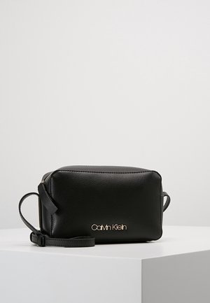 Sac bandoulière en cuir noir Calvin Klein avec logo doré et sangle réglable sur un socle blanc sur un fond uni.