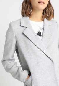Cappotto grigio in misto lana con design doppiopetto, colletti a risvolto e tessuto texturizzato. Presenta maniche leggermente arrotolate e tasche frontali.