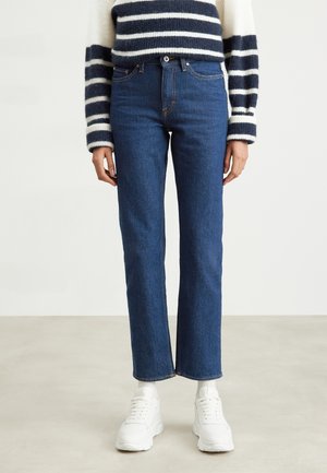 Personne portant un jean droit bleu marine, un pull rayé blanc et bleu marine, des chaussettes blanches et des baskets blanches, debout sur un sol clair.