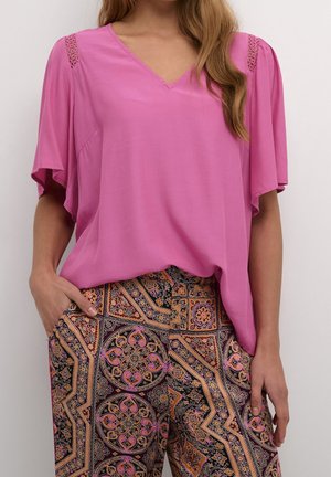 Frau trägt ein locker sitzendes, rosafarbenes Kurzarmoberteil mit Spitzen-Details an den Schultern und gemusterte Hosen mit geometrischen und floralen Designs.