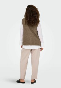 Bruine gebreide vest over een witte lange mouw shirt, gecombineerd met lichtbeige broek en zwarte platte schoenen. Krullend haar zichtbaar.