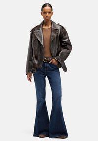 Brązowa skórzana kurtka motocyklowa z pagonami, zestawiona z dopasowanym brązowym topem i rozszerzanymi niebieskimi dżinsami. Gładka faktura i wyraźne szwy.