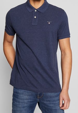 Man wearing a navy blue short-sleeve polo shirt with two buttons and small logo on left chest, paired with medium blue jeans.
