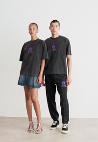 Et par modeller iført matchende mørkegrå oversized t-skjorter med fargerik logo. En modell i en denimnederdel, den andre i mønstrede bukser og sneakers.