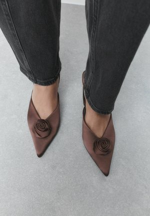 Feet wearing brown pointed-toe shoes with rose decoration, paired with black jeans, standing on a light gray floor.