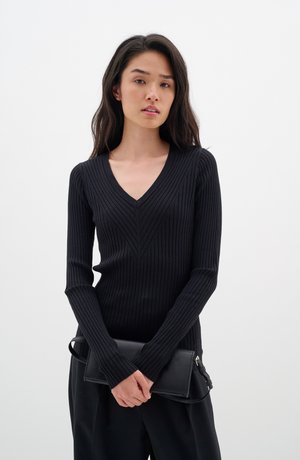 Schwarzes geripptes Langarmshirt mit V-Ausschnitt, ausgestattet mit vertikaler Rippung und einer figurbetonten Silhouette, kombiniert mit einer schwarzen Clutch.
