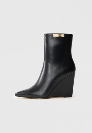 Bottine noire en cuir à semelle compensée avec bout pointu et étiquette de marque dorée près du bord supérieur sur fond clair.