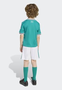 Teal nogometna majica s belimi ovratniki in črtami, v kombinaciji z belimi hlačami in teal nogavicami. Oblika vključuje logo na levem rokavu.