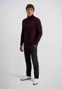 Mann, der mit einer Hand in der Tasche steht, trägt einen dunkelvioletten Rollkragenpullover, schwarze Hosen und weiße Turnschuhe vor einem schlichten Hintergrund.