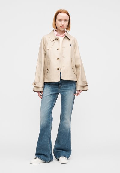 Μπεζ βαμβακερό μπουφάν με γιακά και τέσσερα κουμπιά μπροστά, συνδυασμένο με ανοιχτό μπλε καμπάνα τζιν και λευκά αθλητικά παπούτσια, στέκεται μπροστά σε ένα απλό φόντο.