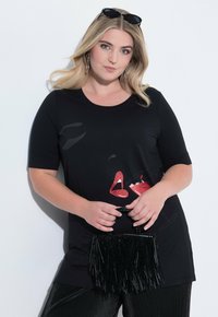 Camisa de manga corta negra con diseños gráficos rojos de labios y un cóctel, acompañada de un bolso de flecos negro. Tela suave con corte relajado.