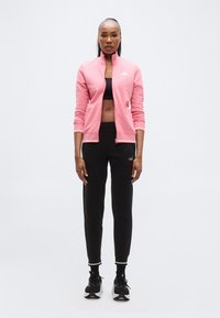 Ροζ φούτερ με φερμουάρ και ριμπ μανσέτες, σε συνδυασμό με μαύρο φόρμας με λευκές λεπτομέρειες και μαύρα αθλητικά παπούτσια.