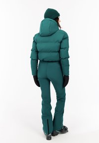 Teal kleurige pufferjack met capuchon, slim fit ontwerp en zwarte handschoenen. Broek in bijpassende teal kleur met ritsen bij de enkels.