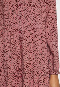 Rosa tygklänning med svart prickig mönster, featuring en knappslå och samlad midja. Långa ärmar med elastiska manschetter.