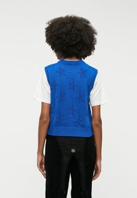 Plavi pleteni bezrukavnik s istaknutim uzorcima zvijezda, rebrastim ovratnikom i rubovima, nosi se preko bijele košulje, uz crne hlače.