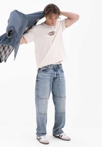 Beżowy t-shirt z logo, jasnoniebieskie dżinsy z naszywkami oraz białe trampki z czarnymi paskami. Mężczyzna trzyma niebieską kurtkę z kratkowaną podszewką.