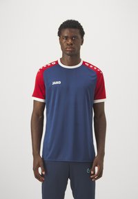 Maglietta sportiva blu navy con maniche a motivo rosso, colletto e bordi bianchi, con il logo JAKO sul davanti.