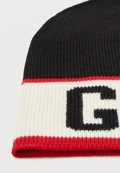 Gorro preto canelado com uma aba de cor creme, apresentando uma letra "G" preta em destaque e uma faixa vermelha ao longo da borda inferior.