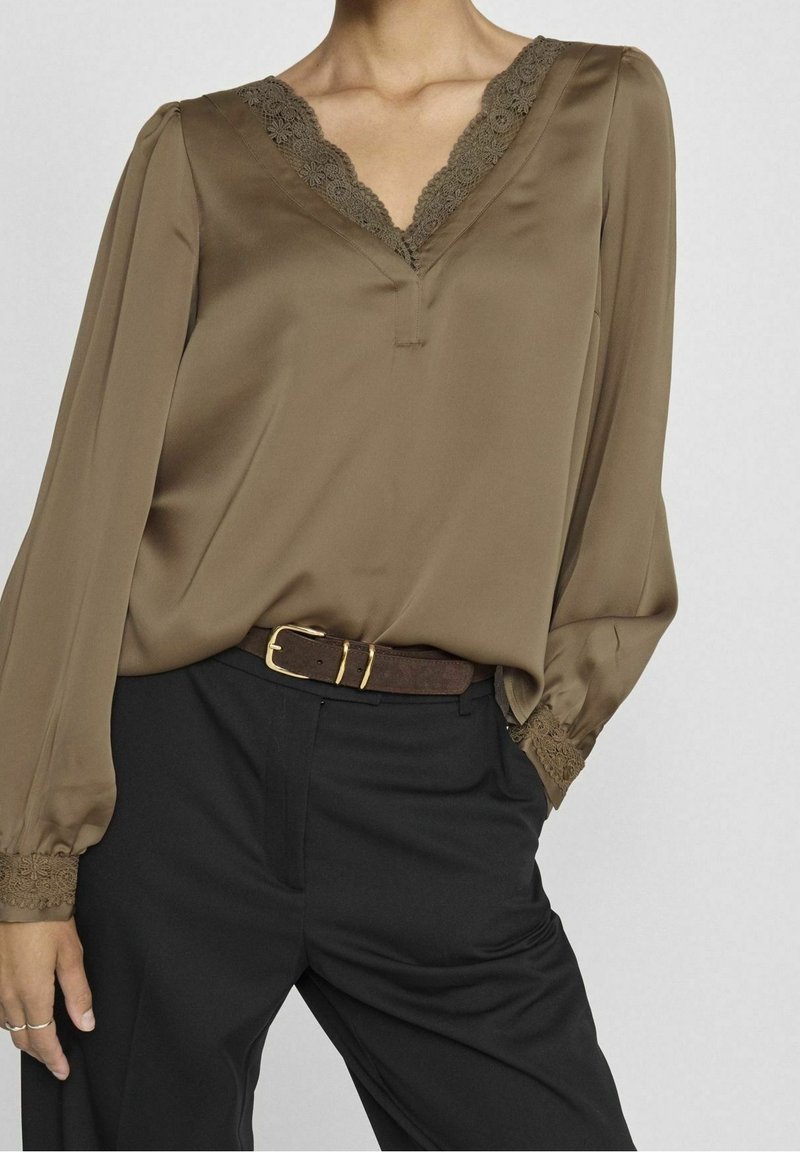 Femme portant un chemisier marron à manches longues avec bordure en dentelle et un pantalon noir, une ceinture marron avec une boucle dorée, main reposant dans la poche.