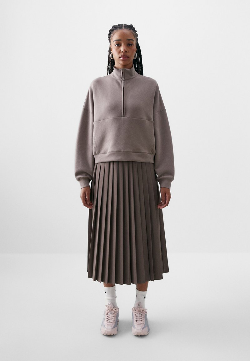 Sweatshirt cinza mesclado com fecho de zíper e um ajuste descontraído, combinado com uma saia midi plissada em um tom semelhante e ténis de desporto rosa claro.