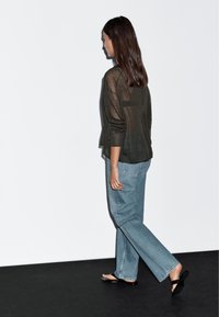 Leichtes, dunkelgrünes Langarmoberteil, kombiniert mit hellblauen, lockeren Jeans und schwarzen flachen Sandalen, von hinten betrachtet.