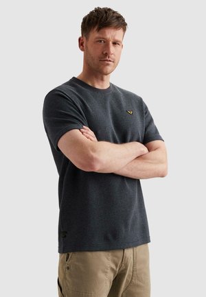 Muž s krátkymi vlasmi oblečený v tmavosivej pruhovanej tričke a béžových nohaviciach stojí so založenými rukami na rovnom pozadí.