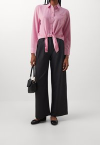 Camisa de manga larga a rayas rosa atada a la cintura, combinada con pantalones de pierna ancha de color gris oscuro. Los accesorios incluyen un pequeño bolso negro y zapatos planos.