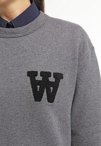 Harmaa huppari, jossa on teksturoitu musta "W" -logo rinnassa. Ominaisuuksina klassinen pyöreä pääntie ja pehmeä, puuvillamainen kangas.