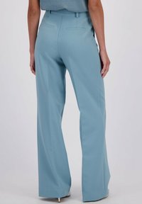 Vrouw in lichtblauwe, hoge taille, wijde broek met achterzakken, staand tegen een lichte achtergrond.