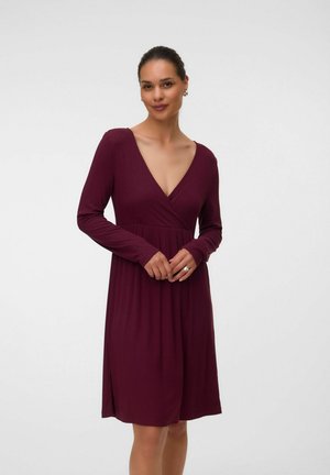 Femme debout portant une robe longue violette foncée à manches longues, décolleté en V profond, longueur au genou, sur fond blanc uni.