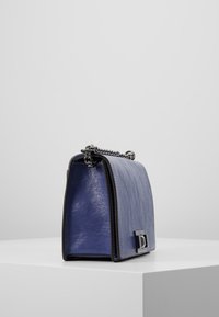 Borsa a tracolla compatta in pelle blu con una forma strutturata, hardware in argento e tracolla a catena. Presenta una finitura testurizzata e un dettaglio di chiusura frontale.