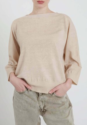 Donna che indossa un maglione beige dalla vestibilità ampia con maniche a tre quarti e jeans grigio chiaro a vita alta, con le mani infilate nelle tasche.