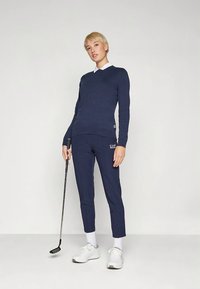 Temno moderen pulover z V-izrezom, kombiniran s temno modrimi ohlapnimi hlačami z blagovno znamko EA7. Model drži golf palico in nosi bele superge.