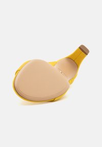 Keltainen mokkanahka korkokenkä, jossa on pyöreä kärki ja sileä beige pohja. Kannan yläosassa on hienovarainen ruskea yksityiskohta.
