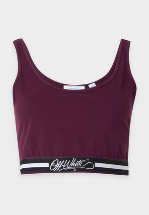 Joven mujer con top deportivo burdeos y leggings, con zapatillas negras y blancas, está de pie con las manos detrás de la espalda sobre un fondo liso.