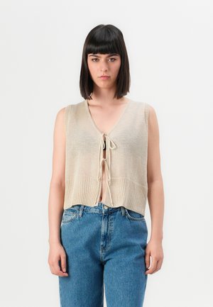 Ung jente med rett svart hår som har på seg en beige ermeløs strikket vest knyttet foran og høy midje blå jeans, som står.