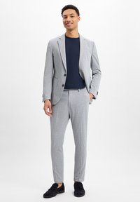 Hellgraue Sakkojacke mit Revers, kombiniert mit einem dunkelblauen T-Shirt und passenden schmal geschnittenen Hosen; abgerundet mit schwarzen Wildlederschuhen.