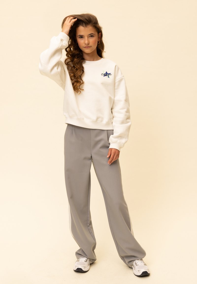 Witte trui met geborduurd sterontwerp, gecombineerd met lichtgrijze loszittende broek. Witte sportschoenen maken de outfit compleet.