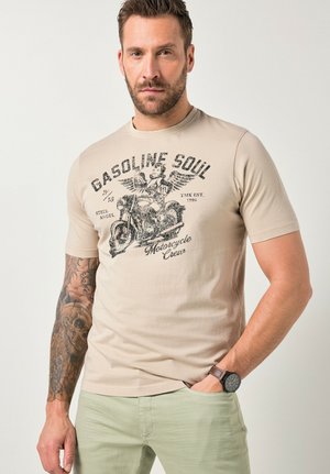 Hombre con brazo tatuado que lleva una camiseta beige con un gráfico de motocicleta y ángel alado, pantalones verde claro y reloj de pulsera, de pie con la mano en el bolsillo.
