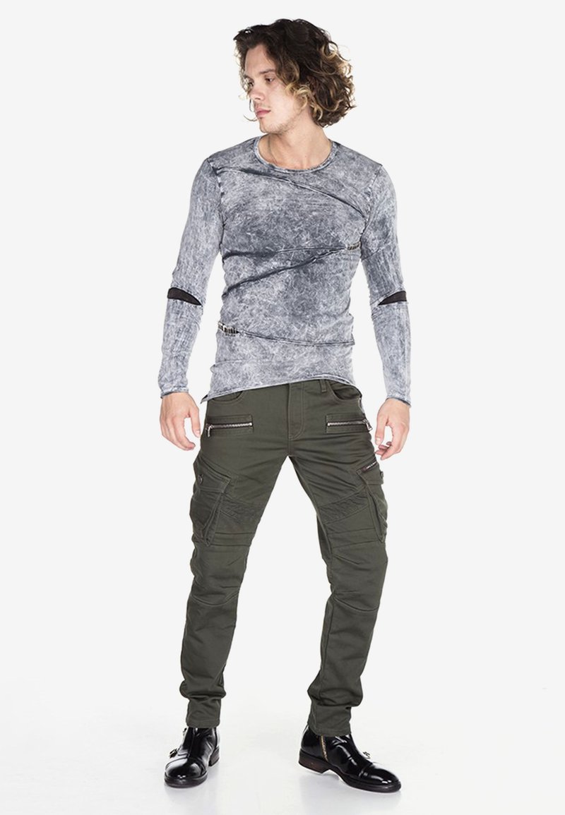 Grijze long-sleeve top met een gestructureerd patroon en asymmetrische stiksels, gecombineerd met donkergroene cargo-panths met zakken en ritsen. Zwarte schoenen.