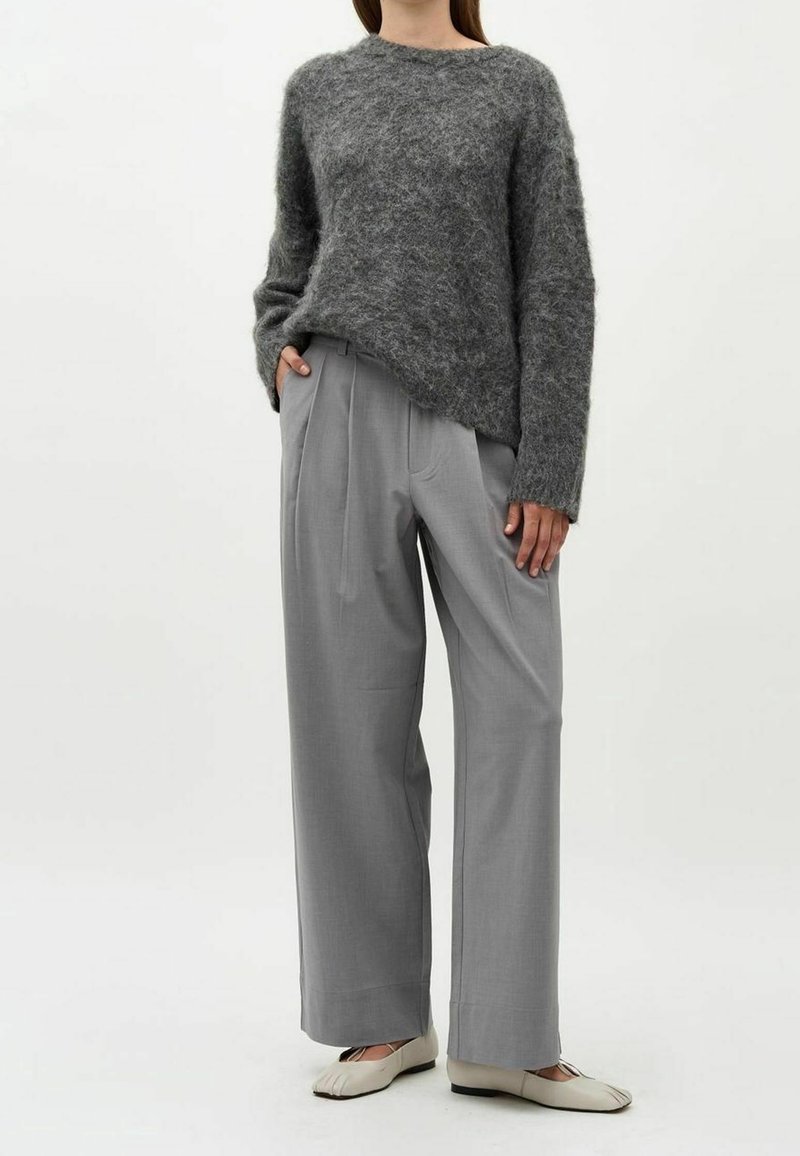 Femme portant un pull texturé gris foncé, un pantalon large gris clair et des ballerines à bout fendu beige clair, debout les mains dans les poches.