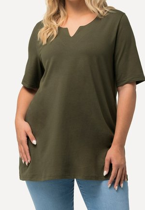 Mujer con una túnica de manga corta de color verde oliva oscuro con escote en forma de muesca y vaqueros azul claro, manos apoyadas cerca de las caderas.
