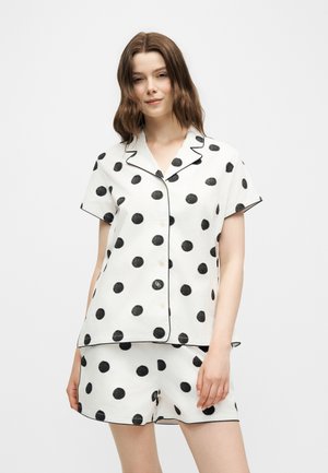 Femme portant un haut blanc à manches courtes boutonné et un short assorti avec de gros pois noirs et un passepoil noir sur le col et les bords.