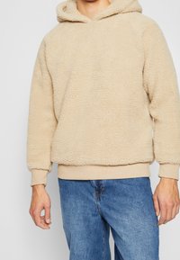 Beige Fleece-Kapuzenpullover mit strukturierter Oberfläche, Kordelzug an der Kapuze und gerippten Bündchen. Kombiniert mit hellblauen Denim-Jeans.