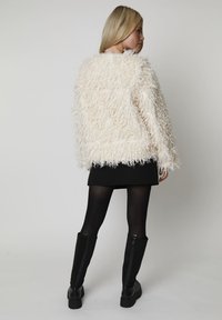 Teksturowana, puszysta kurtka w kremowym kolorze o luźnym kroju, noszona na czarnej mini spódnicy i kryjących czarnych rajstopach. Czarnymi, sięgającymi kolan kozakami dopełniono stylizację.