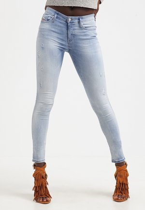 Jasnoniebieskie obcisłe jeansy w stylu distressed noszone z brązowymi sandałami na wysokim obcasie z frędzlami, na stojącej osobie z rękami na biodrach.